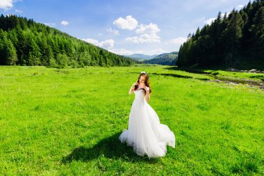 A luxurious bride stands in a meadow in a long white dress and enjoys the amazing landscape.