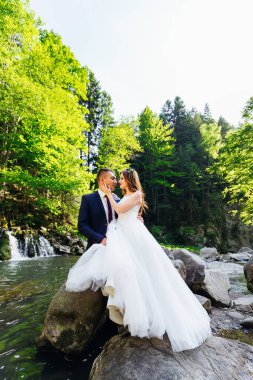 the newlyweds are sitting on a stone near the river in the background green trees.