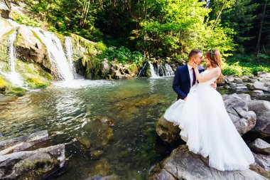 the bride and groom are sitting on a stone in the middle of a mountain river in the forest. Wedding in the forest.