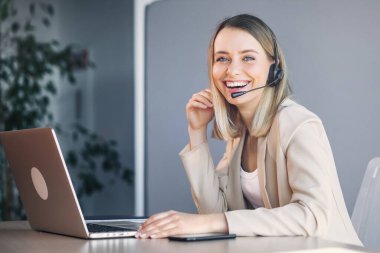 Joyful female call center operator in headset looks at the camera, smiling. Smart business woman sits at the table in the office, works in the support service, advises people