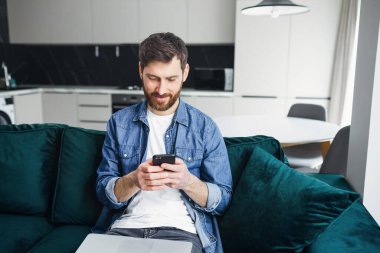 man sitting at home, sending messages on smartphone, smiling, sitting on green sofa in bright living room