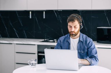 Focused man with wireless headset with microphone looks at computer screen and advises clients on the Internet. A confident tutor gives a private video tutorial.
