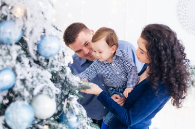 Ebeveynler ve küçük oğulları Noel ağacının yanında. Baba oğluna Noel süslerini gösteriyor..