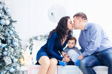 Baba ve anne küçük oğlunu öpüyor. Aile Noel ağacının yakınındaki büyük kutularda hediyelerle oturuyor..