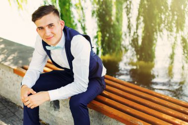 A stylish groom sits on a bench near the lake and looks at the camera