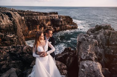 the groom hugs the bride and looks away. rocky shore of the ocean and horizon.
