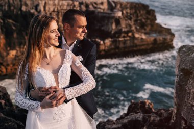 Damat gelini kucaklıyor ve kayaların ve denizin arka planında gülümsüyor. Kapat..