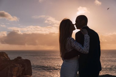 Yeni evliler okyanusun kayalık sahillerinde sarılıp öpüşüyorlar. Gün batımını seyrediyorlar..