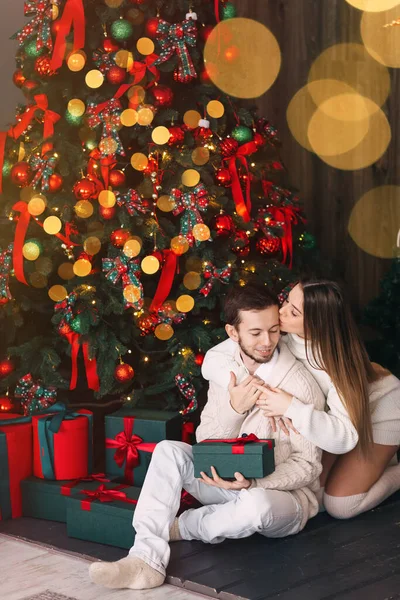 クリスマスツリーの近くに贈り物を持って座っているカップル 男は目を閉じて贈り物を手にし女の子は頬にキスをする — ストック写真