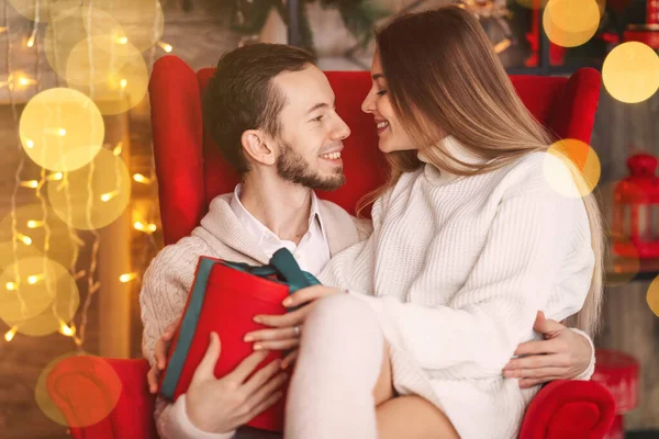 guy and girl holding a gift and want to kiss. couple sitting in red armchair near fireplace with garlands. close up.