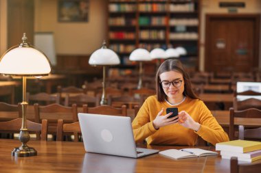 Masada oturan mutlu kız öğrenci genç telefonundaki mesajları okuyor. Kız üniversite kütüphanesinde cep telefonu kullanıyor.