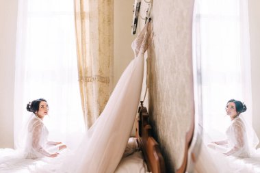 Bornoz ve dantel peçeli güzel bir esmer gelin duvarda asılı bir gelinliğe bakar..