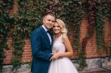 Şık gelin ve damat düğün günlerinde bir yaz parkında duvarın ve sarmaşığın önünde yürürken sarılıp gülümserler.