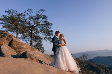 Romantik bir düğün fotoğrafı. Gelin ve damat muhteşem bir gelinlik giymiş. Dağlardaki büyük kayaların yanında sarılıyorlar..