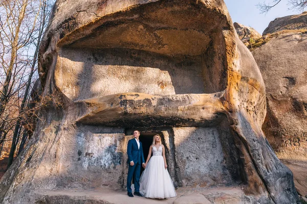 年轻漂亮的新婚夫妇正靠在石墙上 穿着华丽婚纱的新娘 穿着时髦西服的新郎 — 图库照片