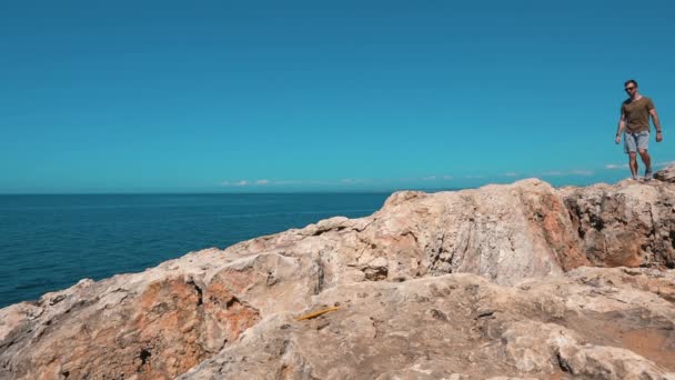 Молодий, спортивний хлопець ходить по скелях, зупиняється і дивиться на море — стокове відео