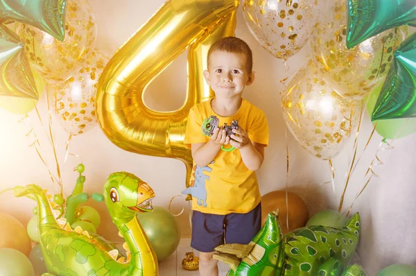 Little boy in a room decorated for a birthday party with golden, green and yellow baloons, large inflatable number 4, presents boxes and ribbons. Photoshooting area at home. Dinosaur baloons.