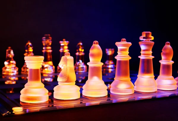 Стеклянные шахматы на шахматной доске, освещенной синим и оранжевым светом — стоковое фото