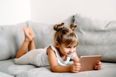 Toddler girl playing with digital wireless tablet computer on couch at home. Baby child growing with online applications. Child and electronic devices concept. Portrait of toddler with smartphone. Education and learning concept.