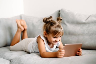 Toddler girl playing with digital wireless tablet computer on couch at home. Baby child growing with online applications. Child and electronic devices concept. Portrait of toddler with smartphone. Education and learning concept.