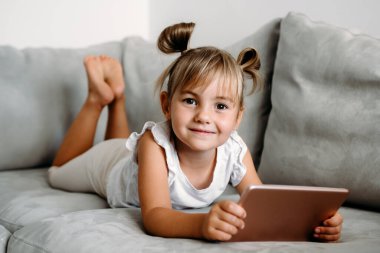 Toddler girl playing with digital wireless tablet computer on couch at home. Baby child growing with online applications. Child and electronic devices concept. Portrait of toddler with smartphone. Education and learning concept.