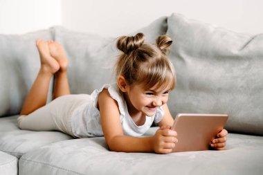 Toddler girl playing with digital wireless tablet computer on couch at home. Baby child growing with online applications. Child and electronic devices concept. Portrait of toddler with smartphone. Education and learning concept.