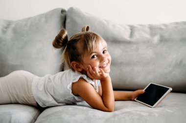 Toddler girl playing with digital wireless tablet computer on couch at home. Baby child growing with online applications. Child and electronic devices concept. Portrait of toddler with smartphone. Education and learning concept.