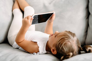 Toddler girl playing with digital wireless tablet computer on couch at home. Baby child growing with online applications. Child and electronic devices concept. Portrait of toddler with smartphone. Education and learning concept.