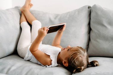 Toddler girl playing with digital wireless tablet computer on couch at home. Baby child growing with online applications. Child and electronic devices concept. Portrait of toddler with smartphone. Education and learning concept.