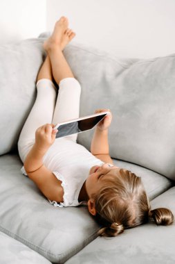 Toddler girl playing with digital wireless tablet computer on couch at home. Baby child growing with online applications. Child and electronic devices concept. Portrait of toddler with smartphone. Education and learning concept.