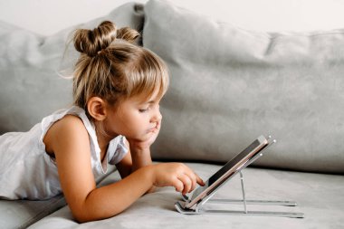 Toddler girl  lies on couch at home and playing with digital wireless tablet computer. Baby child growing with online applications. Child and electronic devices concept. Portrait of toddler with smartphone. Education and learning concept.