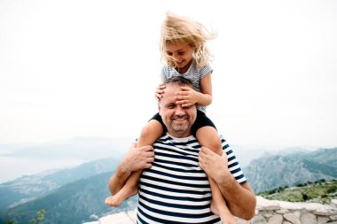 dad and daughter smiling and having fun together. daughter sitting on dad's shoulders and covers dad's eyes with hands. They are standing on the view point on Kotor bay, Montenegro. Wide angle photo