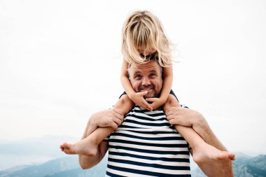 dad and daughter smiling and having fun together. daughter sitting on dad's shoulders and covers dad's eyes with hands. They are standing on the view point on Kotor bay, Montenegro. Wide angle photo