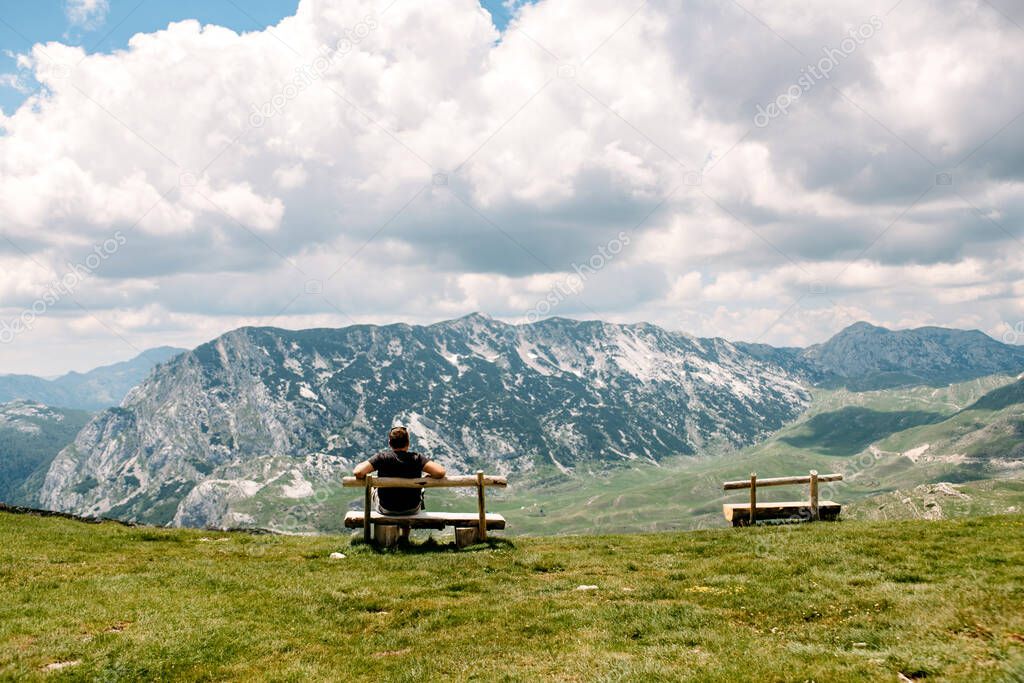 Single millenial man sits on a bench on a mountain plateau and look at the landscape. Traveling alone, loneliness. Human and nature. Zabljak, Montenegro