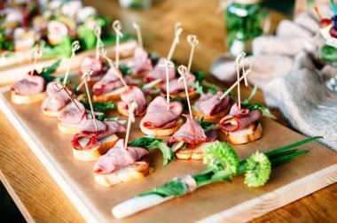 Delicious catering banquet buffet table decorated in rustic style in the garden. Different snacks, sandwiches with ham and greenery on a wooden plate. Outdoor.