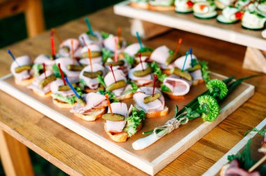 Delicious catering banquet buffet table decorated in rustic style in the garden. Different snacks, sandwiches with ham, cucumber gherkins and greenery on a wooden plate. Outdoor.