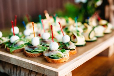 Delicious catering banquet buffet table decorated in rustic style in the garden. Different vegetarian snacks, sandwiches with fish, zucchini, cucumber, pesto sauce, lemon, pepper and greenery on a wooden plate. Outdoor.