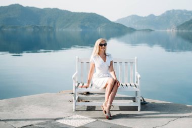 middle aged sexy smiling caucasian woman with sunglasses sits on sea shore in white dress. Summer luxury Europe travel vacation. Mountains  on the background
