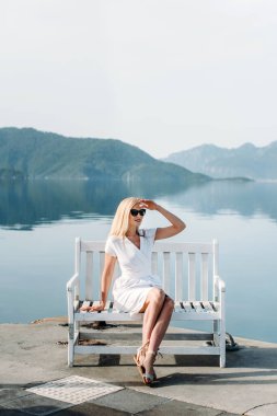 middle aged sexy smiling caucasian woman with sunglasses sits on sea shore in white dress. Summer luxury Europe travel vacation. Mountains  on the background