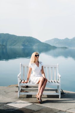 middle aged sexy smiling caucasian woman with sunglasses sits on sea shore in white dress. Summer luxury Europe travel vacation. Mountains  on the background