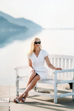 middle aged sexy smiling caucasian woman with sunglasses sits on sea shore in white dress. Summer luxury Europe travel vacation. Mountains  on the background
