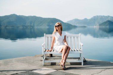 middle aged sexy smiling caucasian woman with sunglasses sits on sea shore in white dress. Summer luxury Europe travel vacation. Mountains  on the background