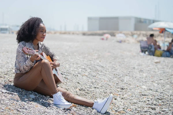 一个快乐的年轻女子坐在岩石沙滩上弹奏着她的吉他 — 图库照片