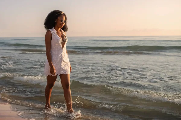 日没時にビーチに沿って歩くサングラス 夏の白いドレスとビキニを持つアフリカの女性 選択的焦点 ストック画像