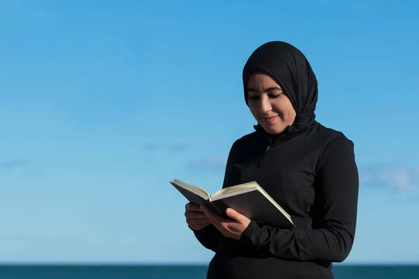 Happy looking muslim woman with a hijab reading a book outdoors on an out of focus background. Selective focus. Diversity and leisure concept.