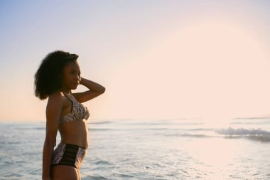 Güneşin batışında plajda bikinili rahat poz veren Afrikalı bir kadının yan görüntüsü. Seçici odak.