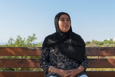 Faslı bir kadının, odaklanamadığı arka planda, tahta bir bankta sessizce oturması. Seçici odaklanma. Boş zaman ve rahatlama konsepti.