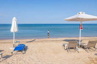 Sakin deniz suyunda duran ve beyaz kumda ufka bakan güzel bir kadın manzarası Yunanistan plajı ve güneşli yataklar şemsiyeleri. Yunanistan. 