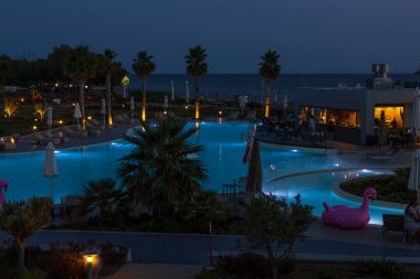 Otele bakan güzel bir gece manzarası. Işıklar, ağaçlar, yüzme havuzu, gece gökyüzüne karşı modern binalar. Yunanistan. Nea Potidea. 03.09.2021.