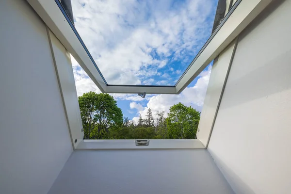 View from dormer roof window in house to tops of forest trees against background of blue sky with white clouds. Sweden.